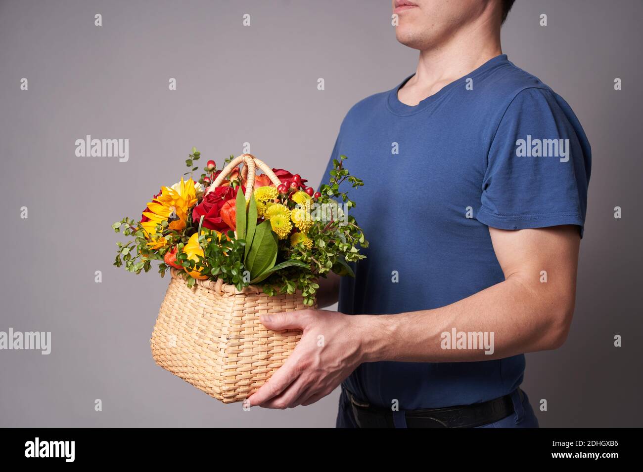 Ein weißer Mann in einem blauen T-Shirt hält einen Blumenkorb mit einem Kopierplatz auf einem grauen Hintergrund aus, Blumenlieferung Stockfoto