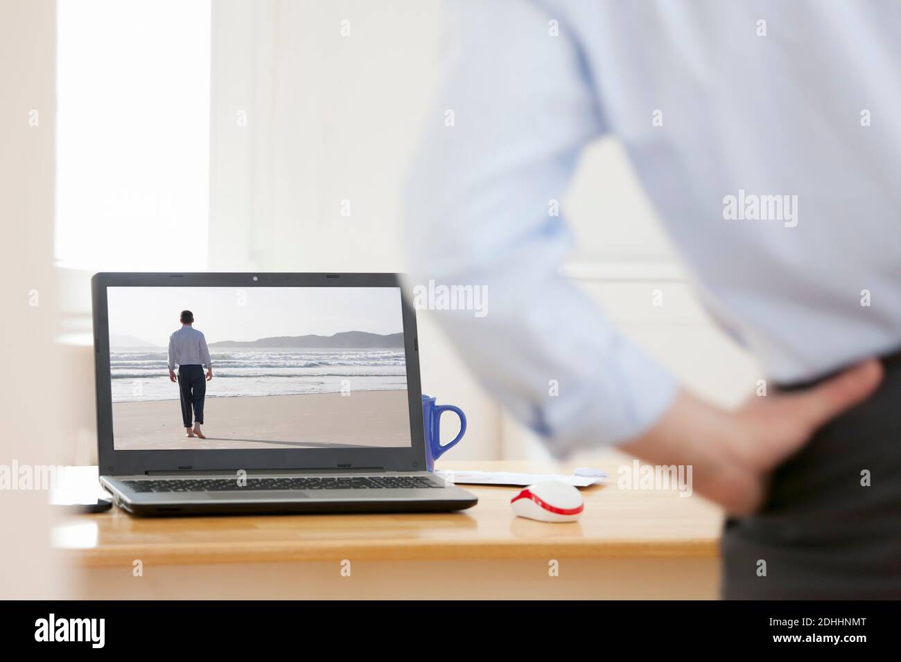 Geschäftsmann, der sich zu Hause an seinem Arbeitsplatz entspannt und wehmütig aussieht Auf einem Laptop mit Geschäftsmann zu Fuß auf einem Strand - Dasselbe Modell auf dem Bildschirm und in Stockfoto