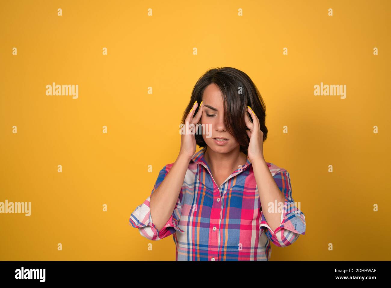 Eine Kopfschmerz habende Frau, die den Kopf hält, der versucht, Migräne zu behandeln, nach unten schaut, während sie gegen die gelbe Wand im Hintergrund mit Kopierraum steht Stockfoto