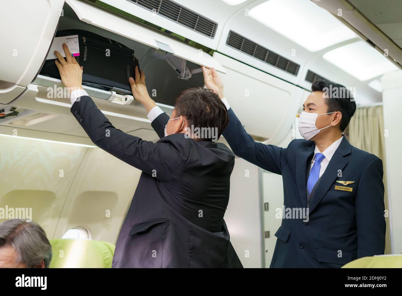 Asiatischer freundlicher Flugbegleiter Mann mit Maske hilft Passagier, Gepäckraum im Flugzeug vor dem Start zu setzen. Stockfoto