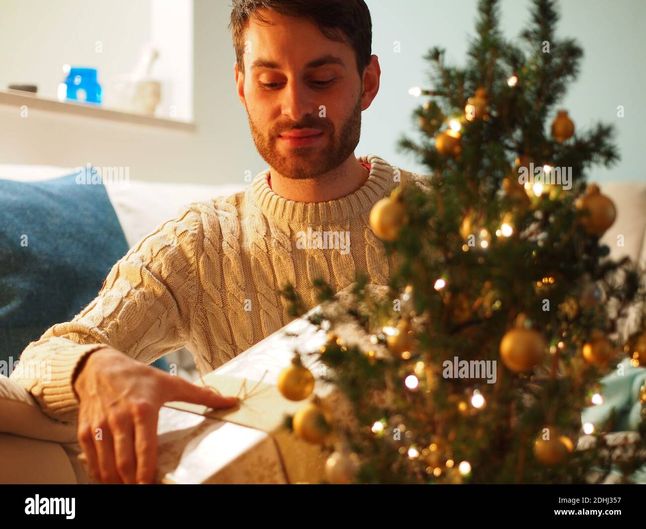 Kaukasischer junger Mann neben einem Weihnachtsbaum in einem Wollpullover, der ein Geschenk in den Händen hält. Er genießt die Ferien in einem gemütlichen Zuhause. Stockfoto