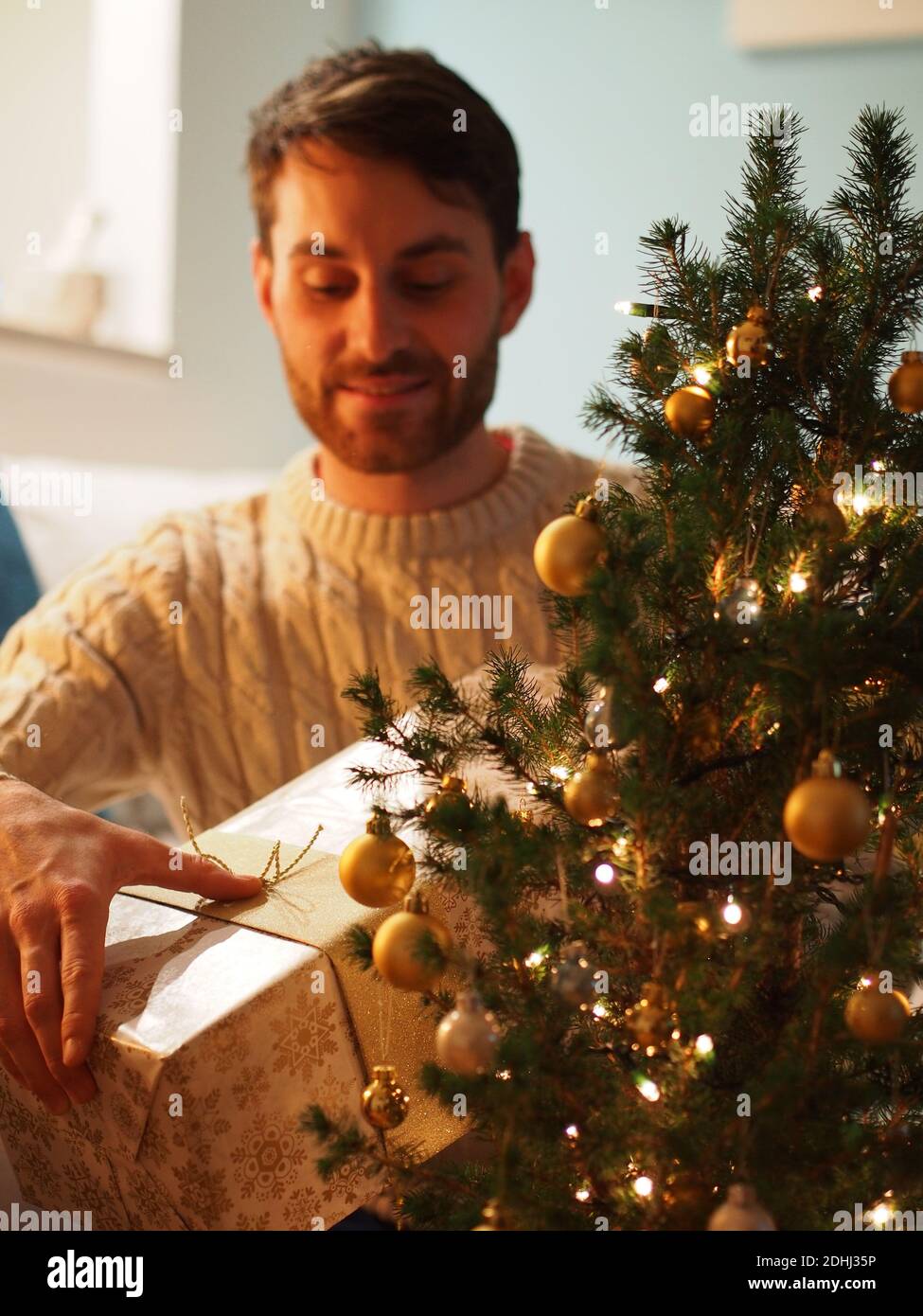 Kaukasischer junger Mann neben einem Weihnachtsbaum in einem Wollpullover, der ein Geschenk in den Händen hält. Er genießt die Ferien in einem gemütlichen Zuhause. Stockfoto