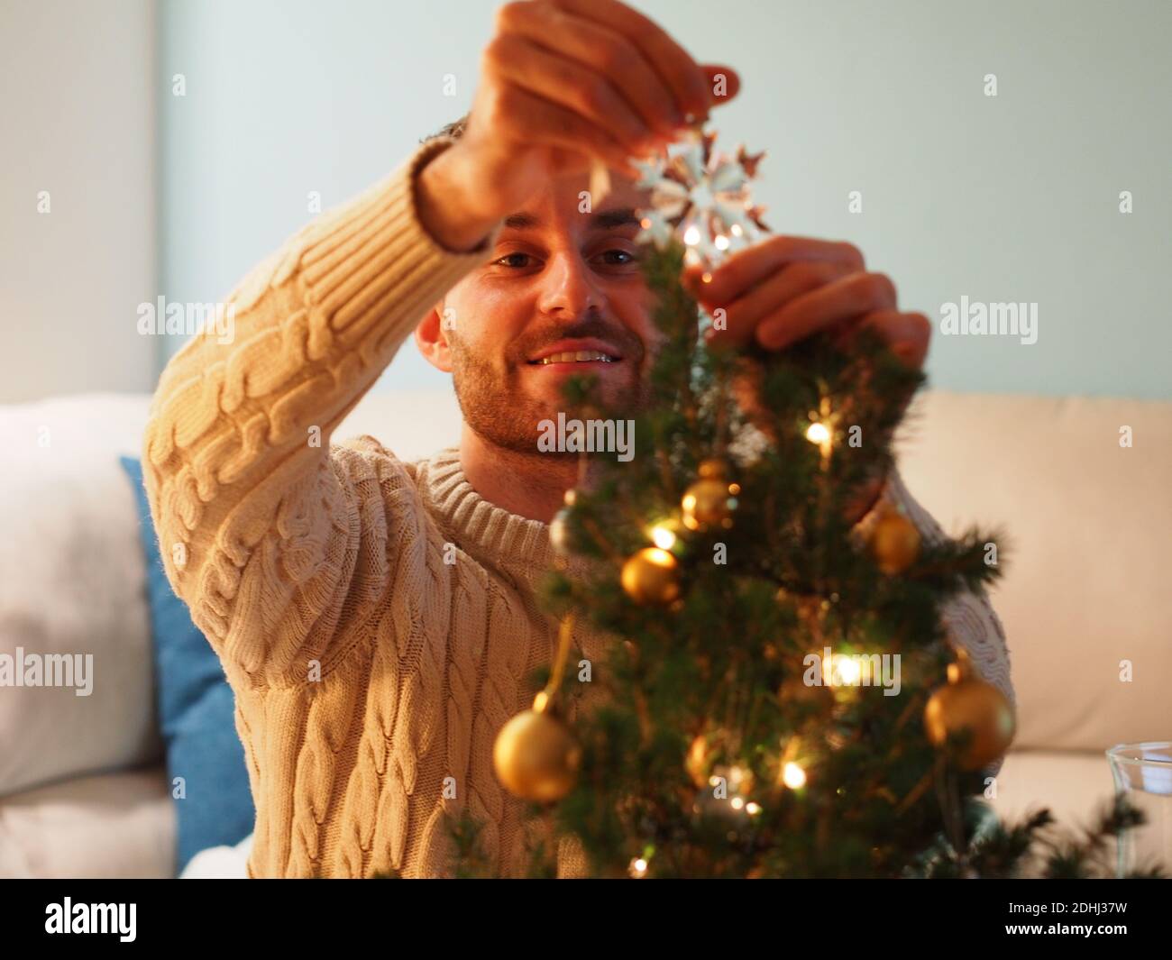 Kaukasischer junger Mann neben einem Weihnachtsbaum in einem Wollpullover, der ein Geschenk in den Händen hält. Er genießt die Ferien in einem gemütlichen Zuhause. Stockfoto