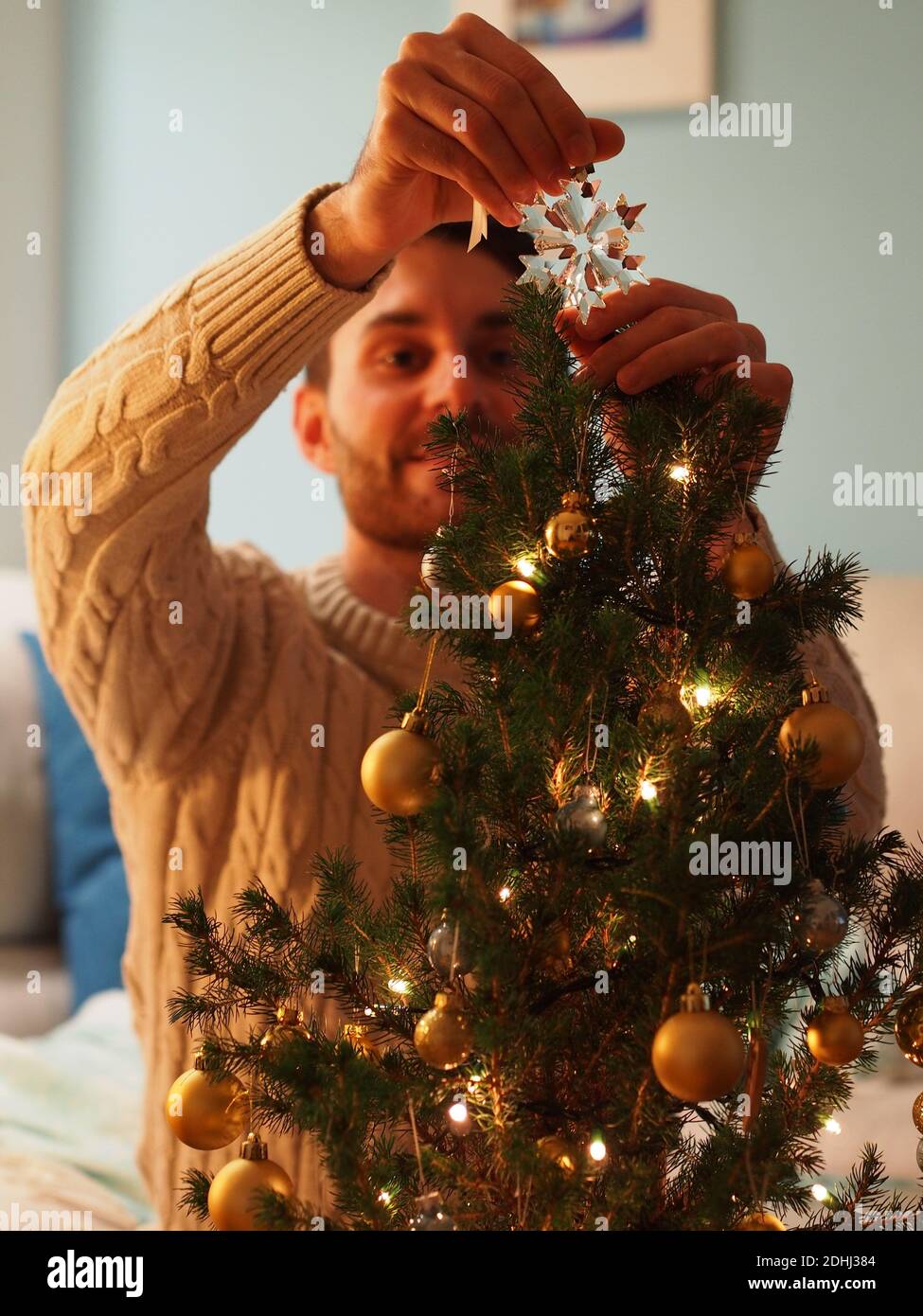 Kaukasischer junger Mann neben einem Weihnachtsbaum in einem Wollpullover, der ein Geschenk in den Händen hält. Er genießt die Ferien in einem gemütlichen Zuhause. Stockfoto