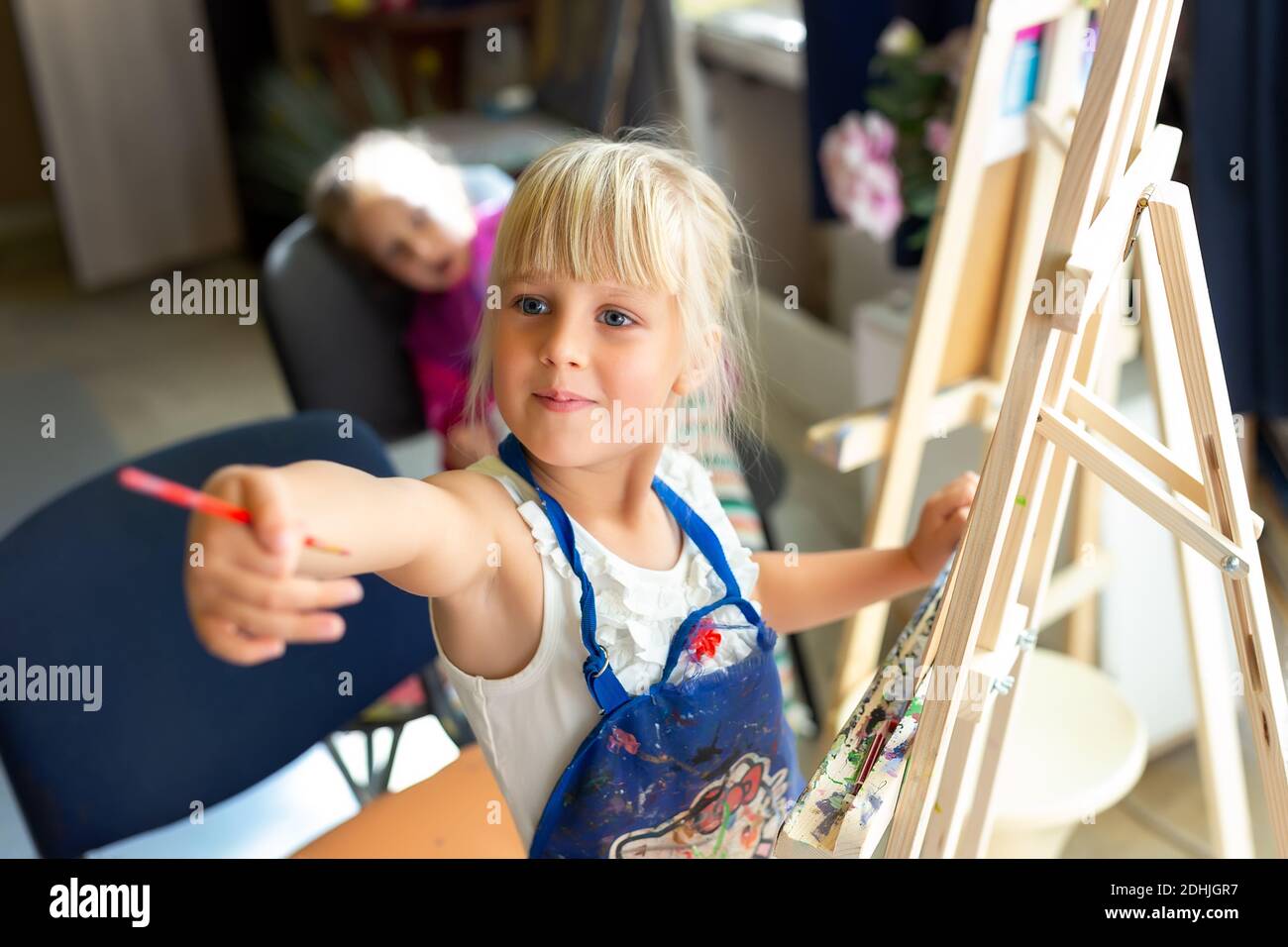 Cute blonde lächelnd kaukasischen Kind Malerei auf Holz Staffelei in der Klasse Workshop Lektion im Kunststudio. Kleines Mädchen hält Pinsel in der Hand und Spaß haben Stockfoto