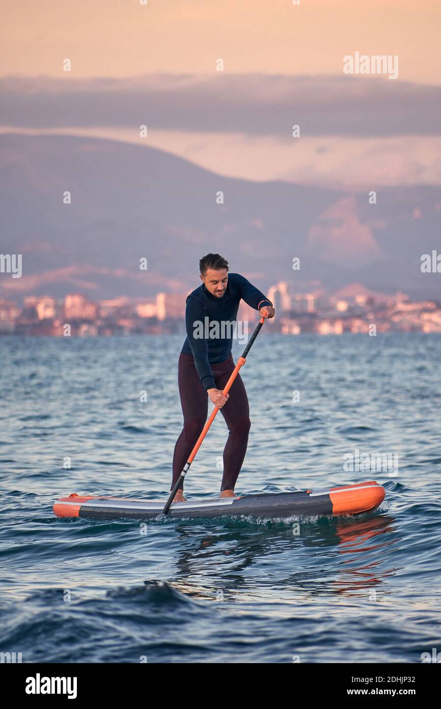 Männlicher Surfer im Neoprenanzug Rudern auf dem Paddelbrett auf dem Meer Wasser auf dem Hintergrund der Berge bei Sonnenuntergang Stockfoto