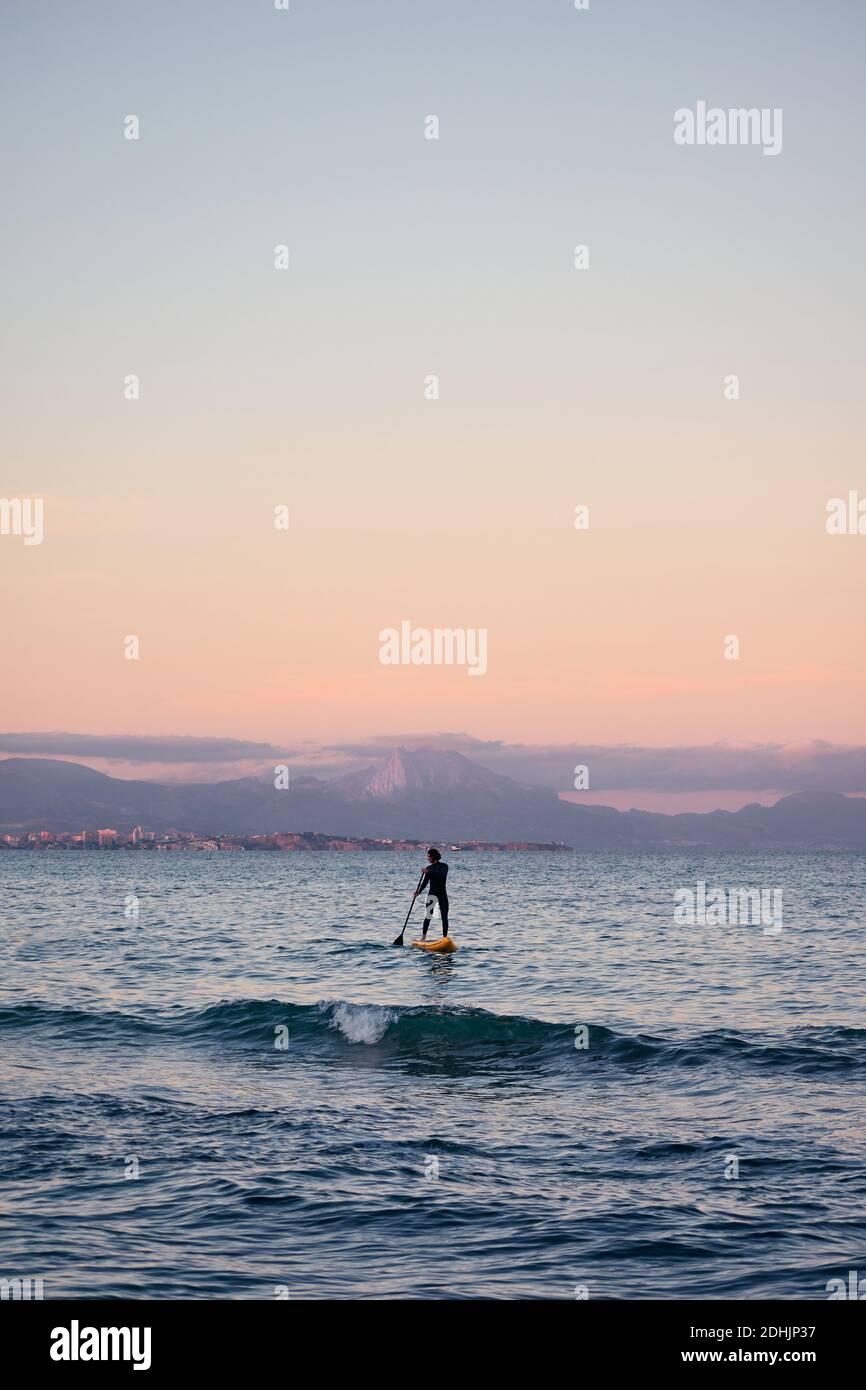 Männlicher Surfer im Neoprenanzug Rudern auf dem Paddelbrett auf dem Meer Wasser auf dem Hintergrund der Berge bei Sonnenuntergang Stockfoto