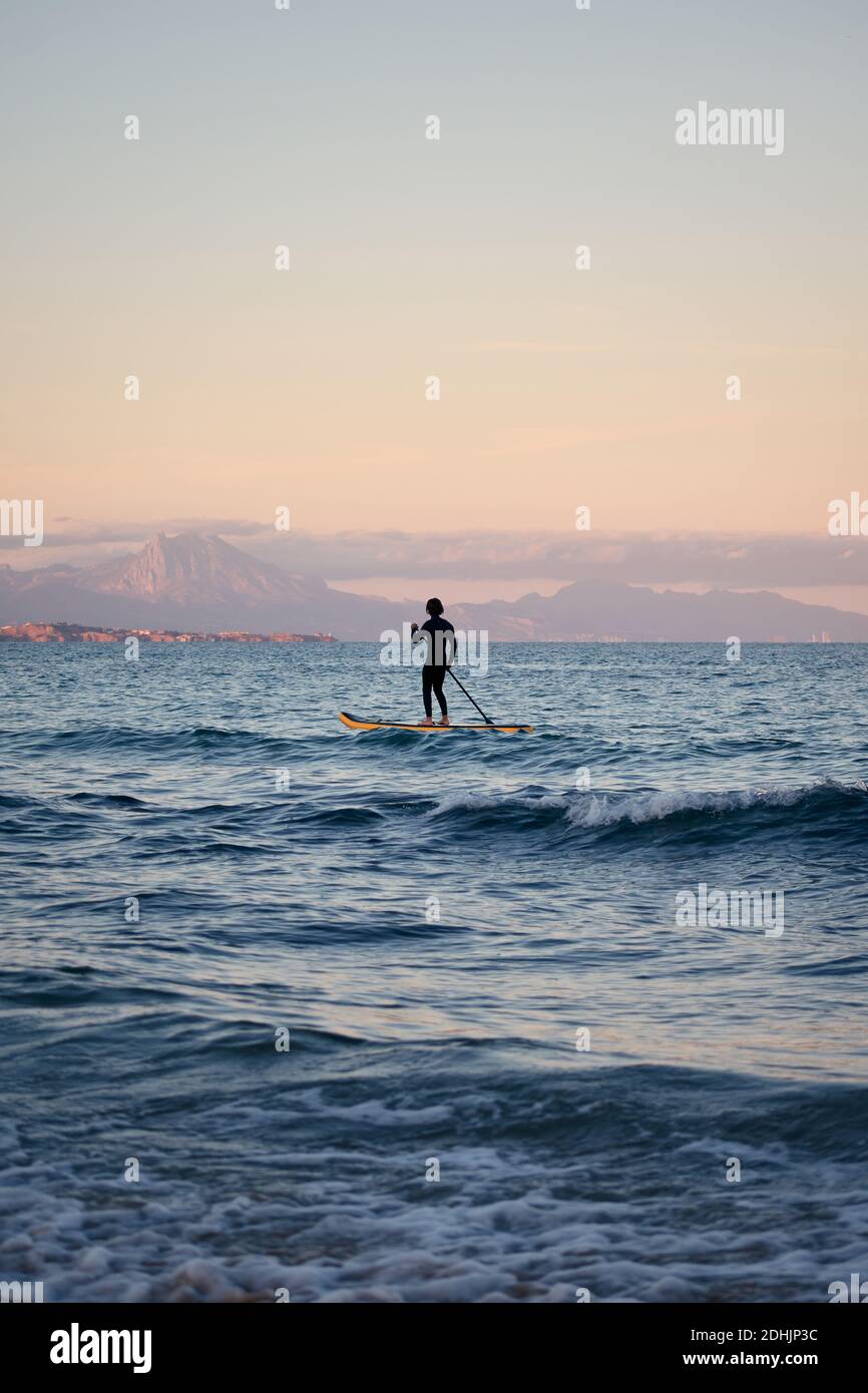Männlicher Surfer im Neoprenanzug Rudern auf dem Paddelbrett auf dem Meer Wasser auf dem Hintergrund der Berge bei Sonnenuntergang Stockfoto