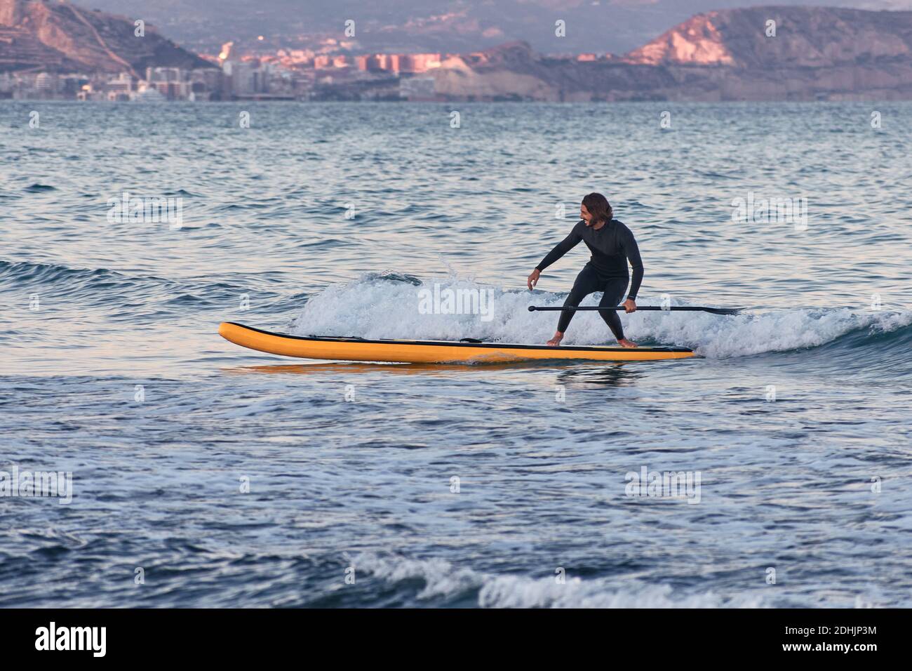 Männlicher Surfer im Neoprenanzug Rudern auf dem Paddelbrett auf dem Meer Wasser auf dem Hintergrund der Berge bei Sonnenuntergang Stockfoto