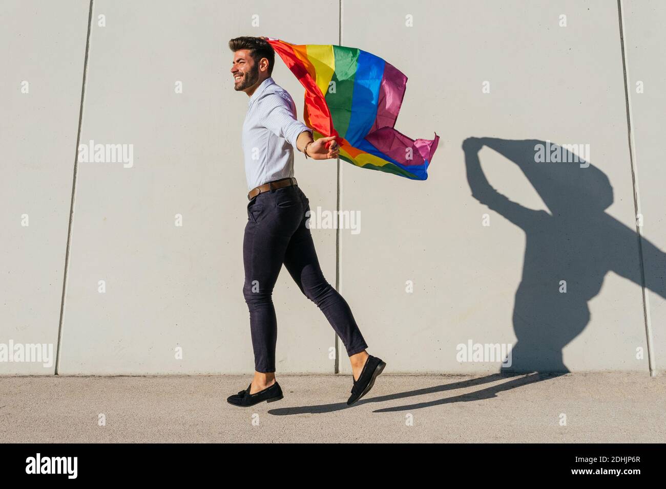 Seitenansicht der fröhlichen homosexuellen männlichen im Moment des Sprüngens Mit Regenbogen-LGBT-Flagge auf der Stadtstraße an sonnigen Tagen Stockfoto