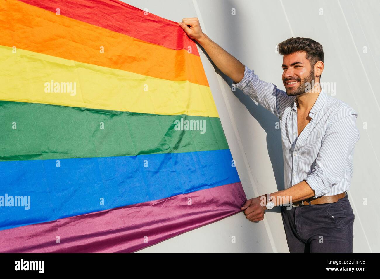 Seitenansicht des fröhlichen homosexuellen Mannes mit LGBT-Flagge in Regenbogen Stehen auf Betonmauer auf der Stadtstraße an sonnigen Tag Stockfoto