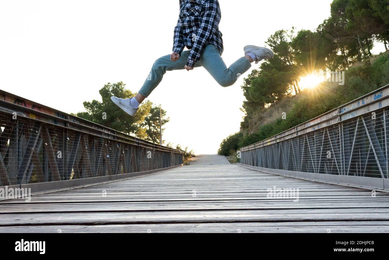 Niedrige Winkel der Ernte anonymen Mann in karierten Hemd und Jeans mit Sneakers, die Spaß haben und hoch über Holz springen Fußgängerbrücke im sonnigen Sommertag Stockfoto