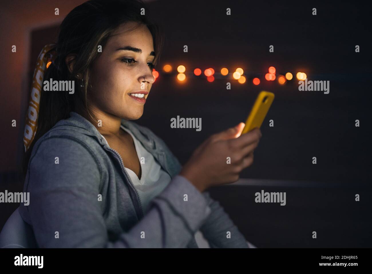 Seitenansicht der glücklichen Frau spielen Videospiel auf Handy Telefon während der Unterhaltung am Abend zu Hause im dunklen Zimmer Stockfoto