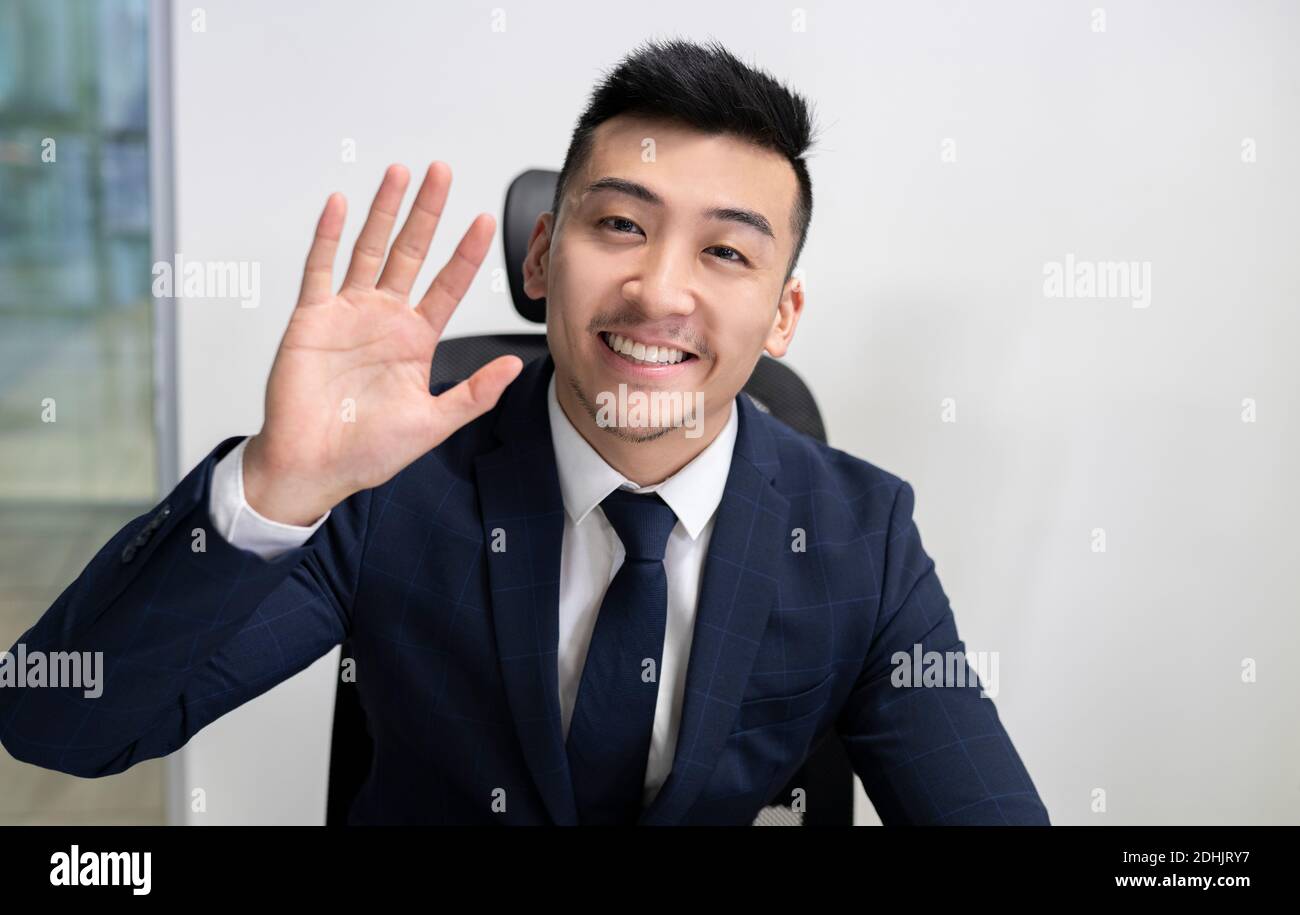 Positive junge asiatische männliche Executive Manager in Business-Stil tragen Blick auf die Kamera und winkende Hand, während Sie im Büro sitzen Stockfoto