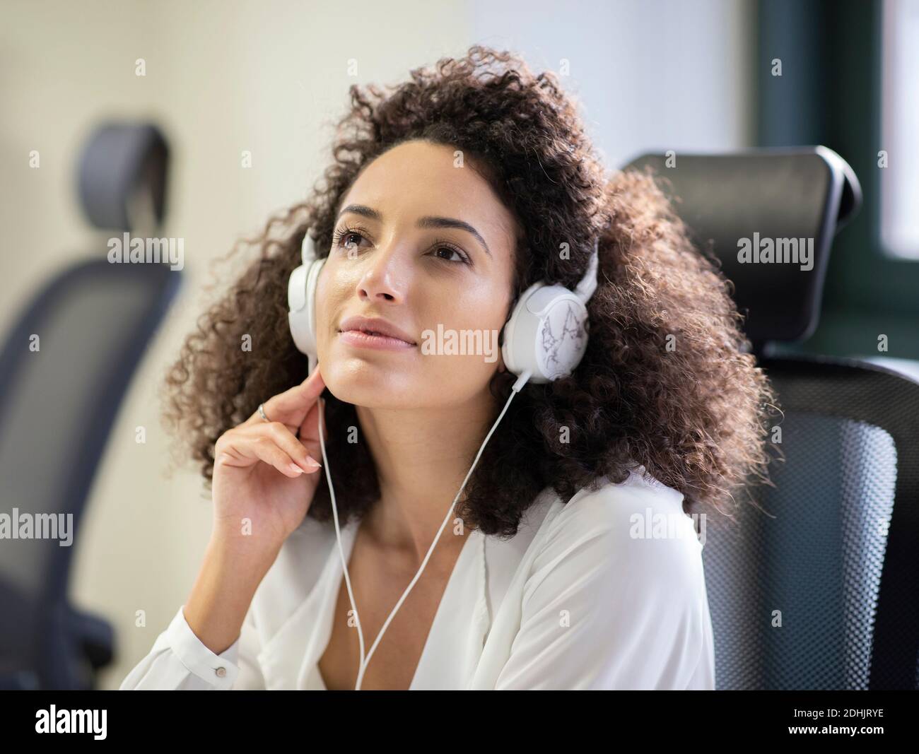 Junge nachdenkliche hispanische Mitarbeiterin in weißer Bluse und Kopfhörern Wegschauen beim Arbeiten am Tisch mit Computer in modernen Arbeitsplatz Stockfoto
