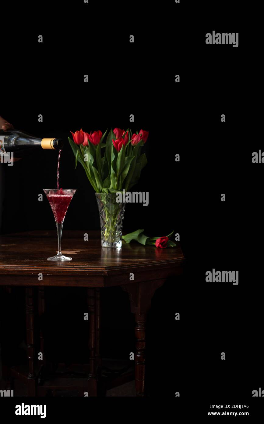Unkenntlich Person mit Flasche Poring Sekt in Glas platziert Auf dem Tisch mit Bouquet von roten Tulpen auf schwarzem Hintergrund Stockfoto