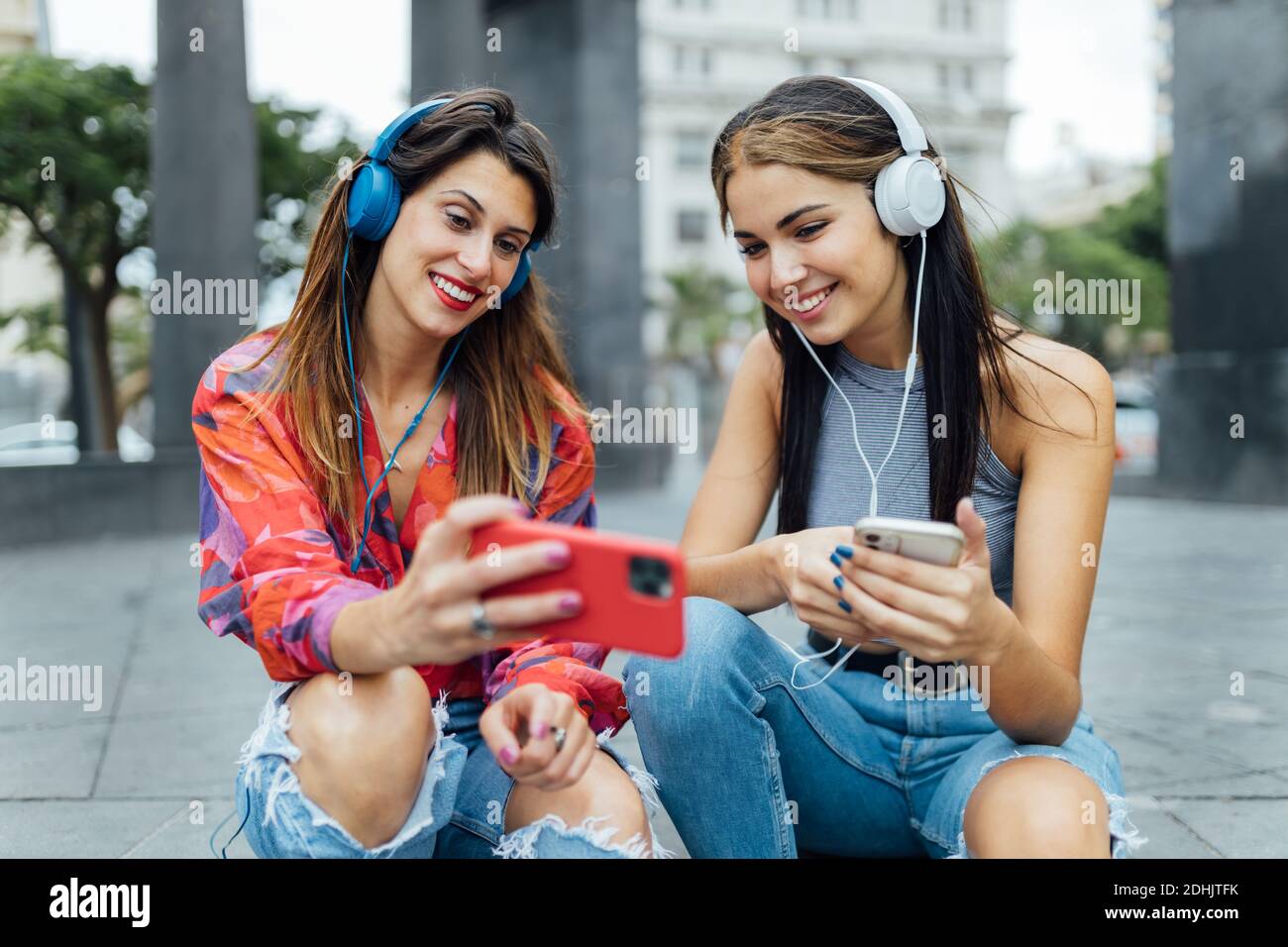 Fröhliche Hipster-Studentinnen in trendigen Outfits surfen in sozialen Netzwerken Auf Mobiltelefonen und Musik mit Kopfhörern hören, während Zusammen sitzen Stockfoto