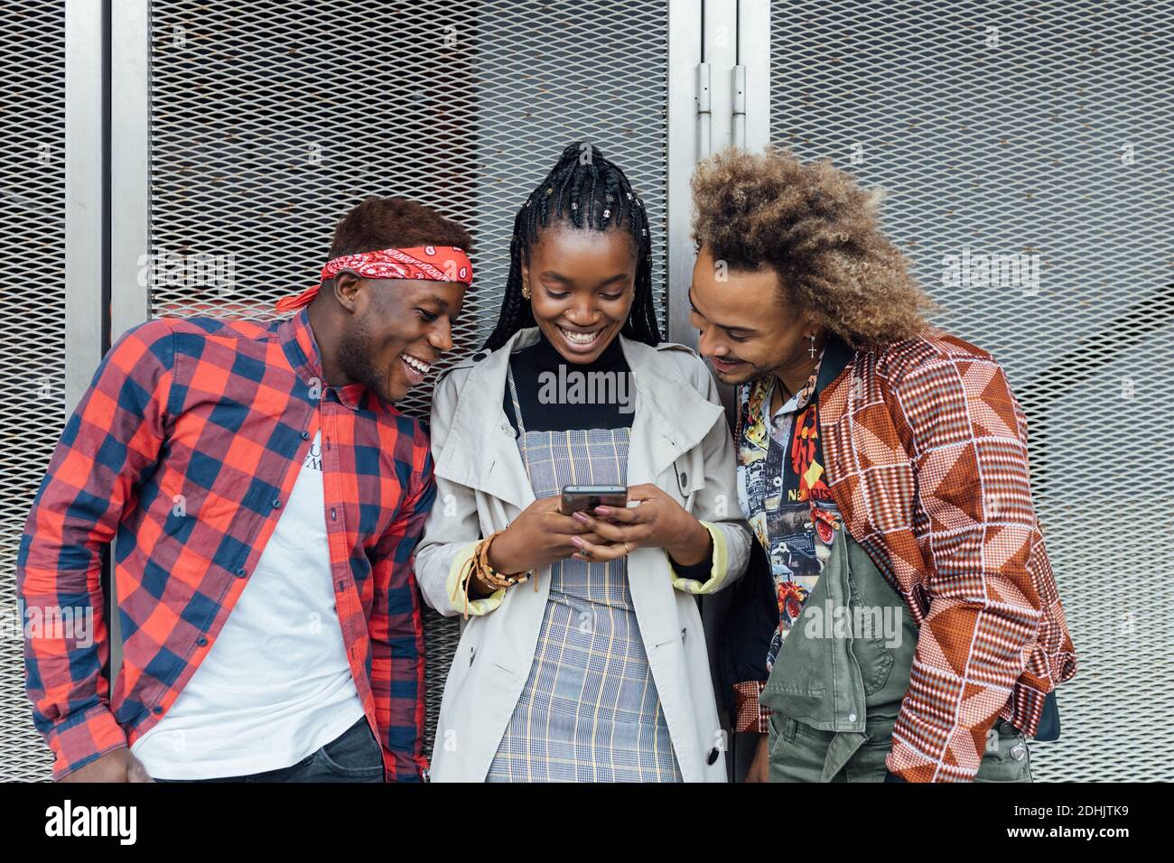 Fröhliche, tausendjährige afroamerikanische Frau und Männer in trendigen Outfits, die an der Wand stehen und Mobiltelefone durchsuchen Stockfoto