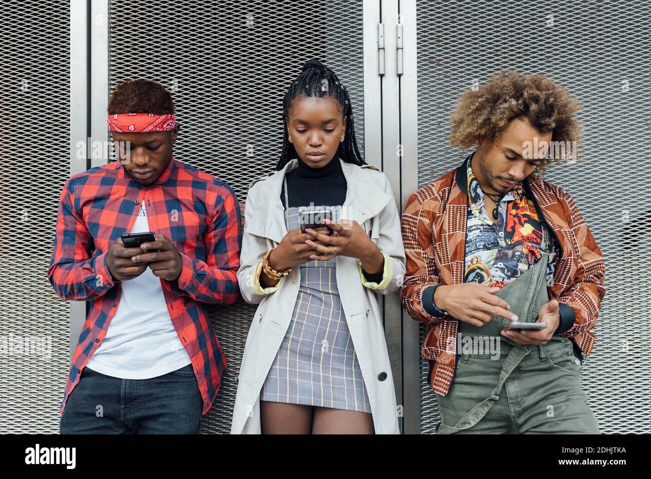 Fokussierte tausendjährige afroamerikanische Frauen und Männer in trendigen Outfits, die an der Wand stehen und Mobiltelefone durchsuchen Stockfoto