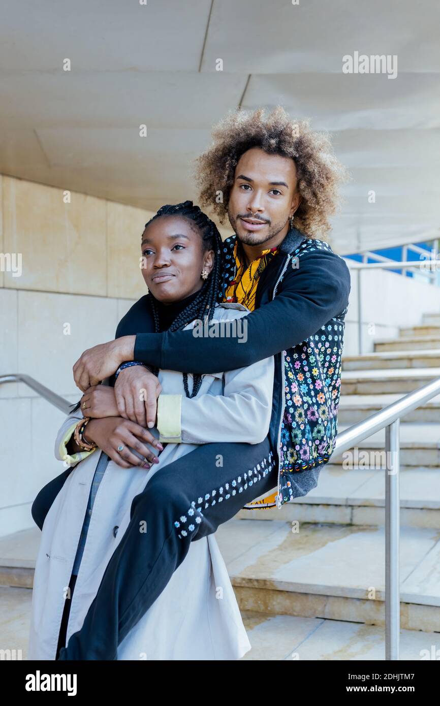 Glücklich junge afroamerikanische Mann umarmt glücklich Freundin während sitzen An der Treppengeländer in der Nähe von modernen Gebäuden während der romantischen Datum in Stadt Stockfoto