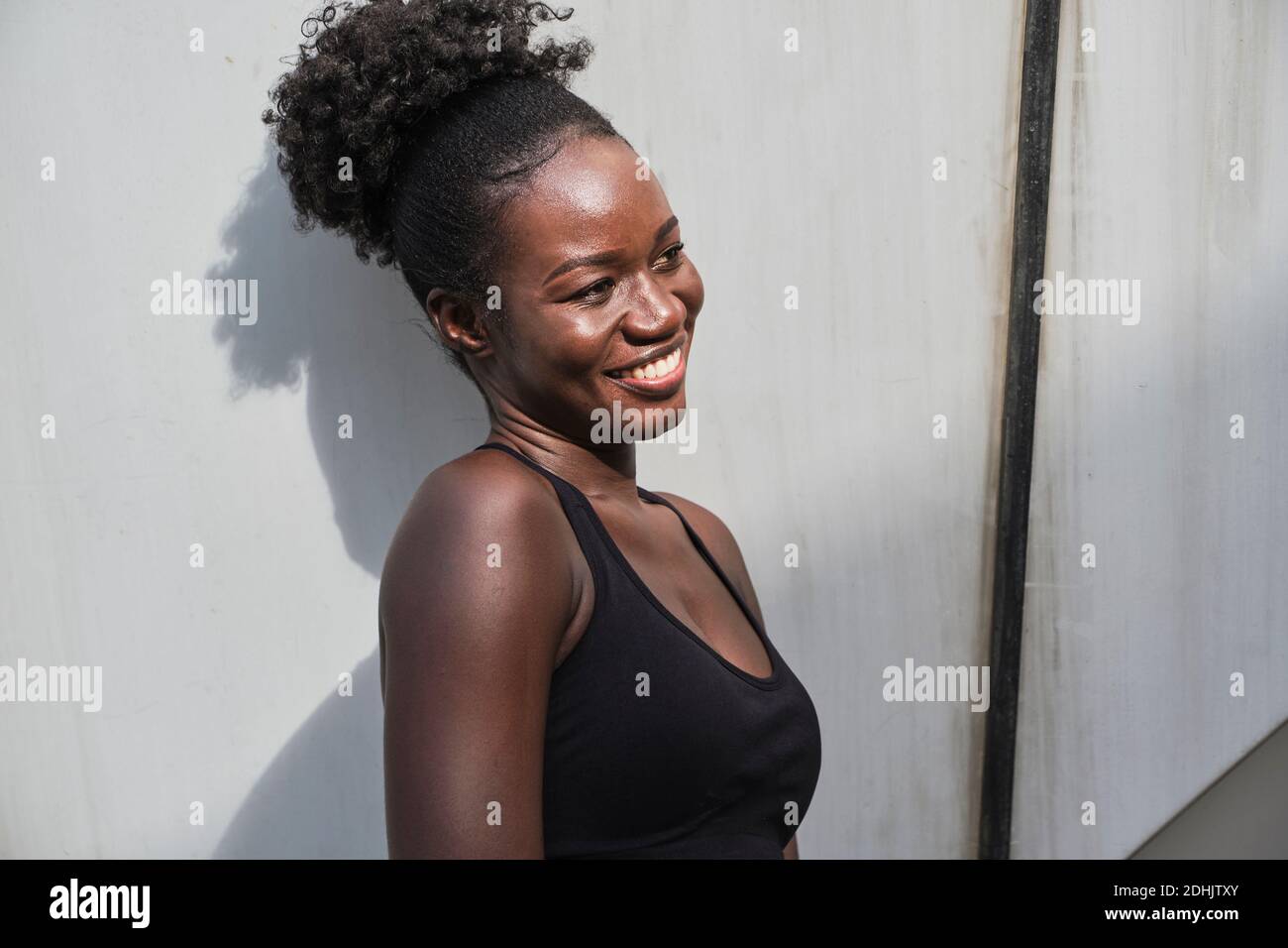 Fröhliche afroamerikanische Athletin trägt Sport-BH lehnt auf Steinmauer des Gebäudes und Blick weg Stockfoto
