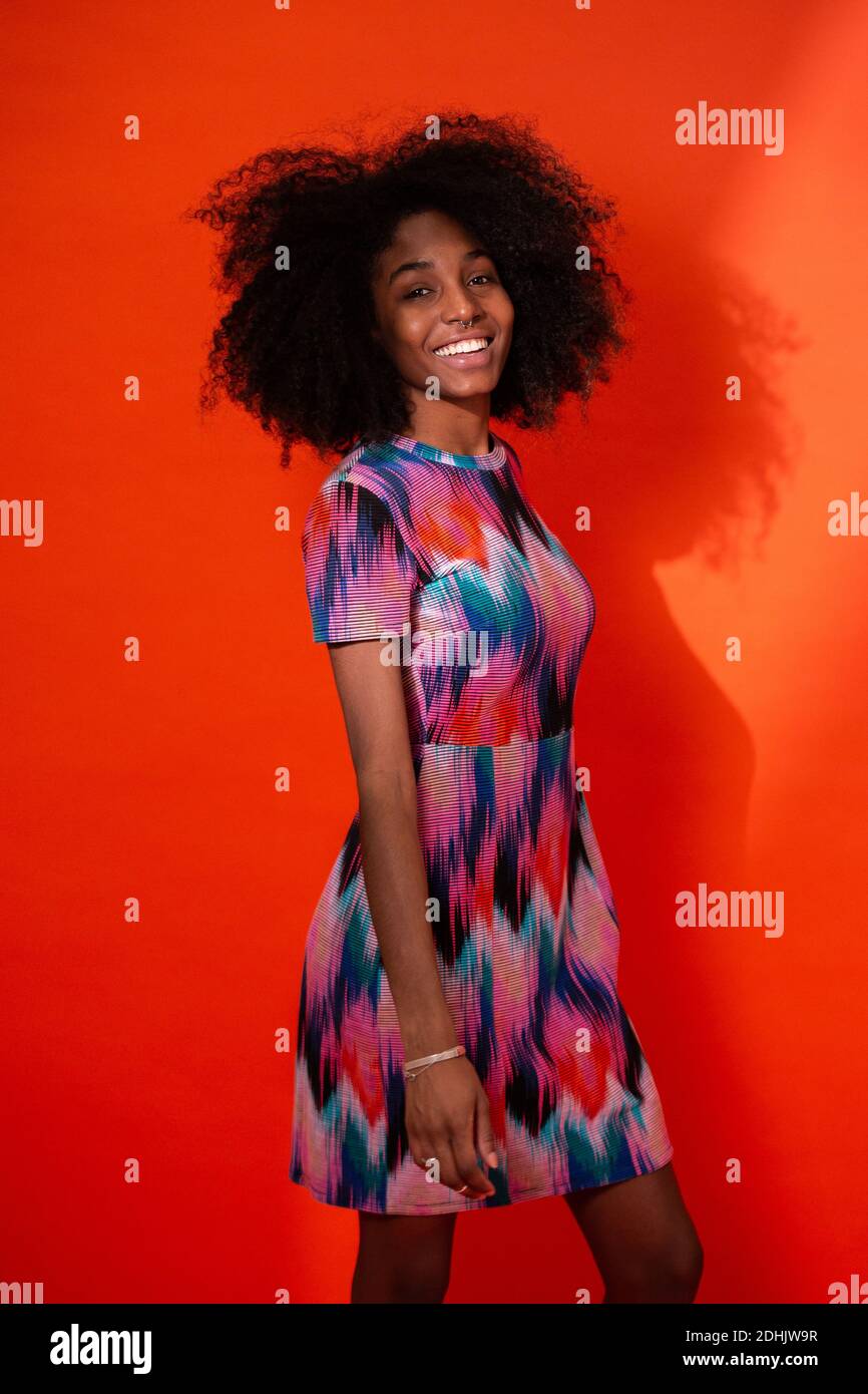 Moderne kubanische Frau mit Afro-Haaren in einem hellen Kleid und lächelnd vor rotem Studiohintergrund Stockfoto