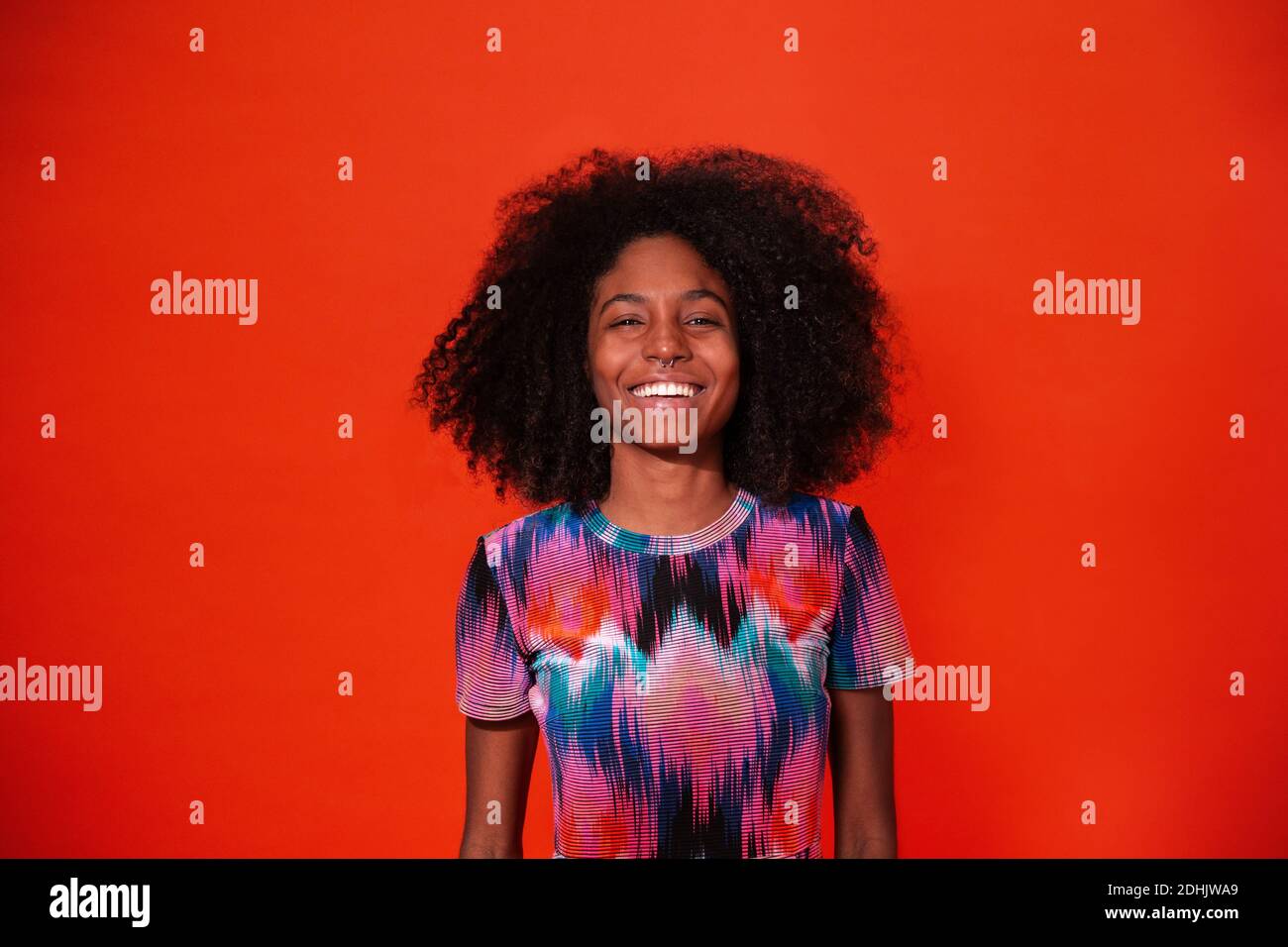 Moderne kubanische Frau mit Afro-Haaren in einem hellen Kleid und lächelnd vor rotem Studiohintergrund Stockfoto