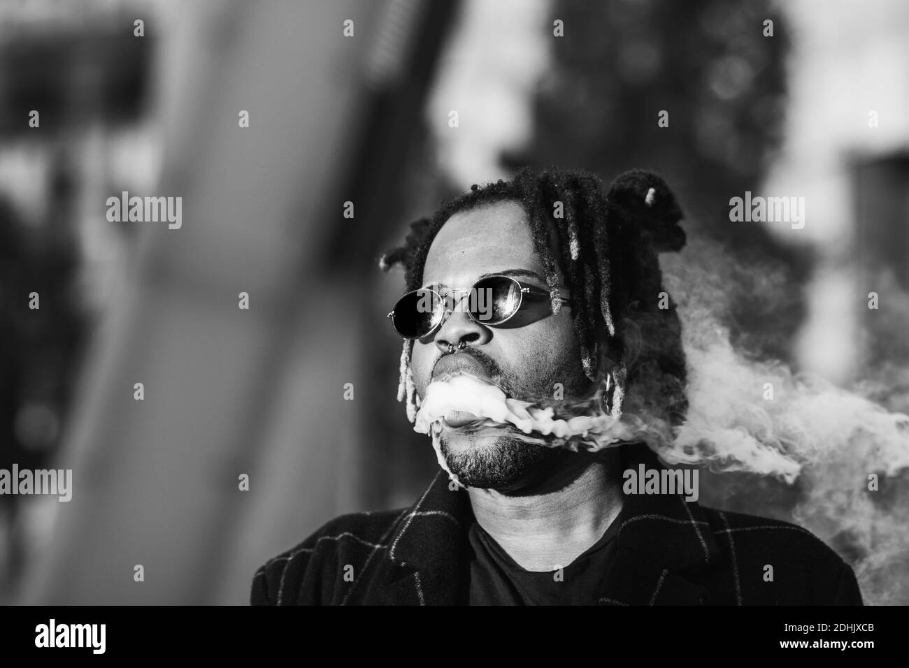Ethnischer Hintermann mit Dreadlocks und Sonnenbrille in dichter Wolke Von Rauch rauchen auf der Straße in der Stadt, die weg schaut Stockfoto