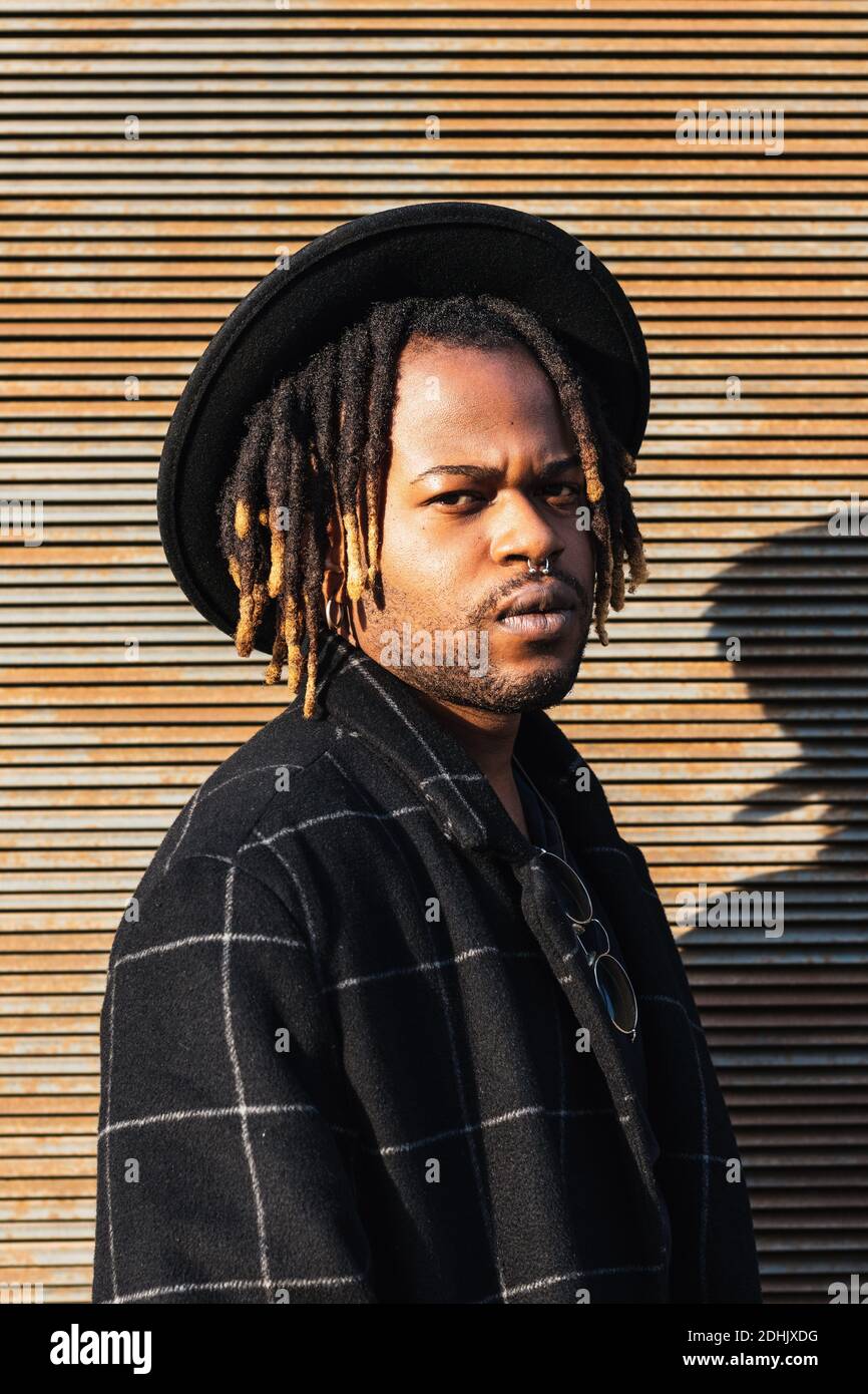 Moderne Erwachsene schwarzen Mann mit Dreadlocks tragen Hut und stilvoll  Mantel Blick auf die Kamera gegen die Straßenwand im Sonnenlicht  Stockfotografie - Alamy