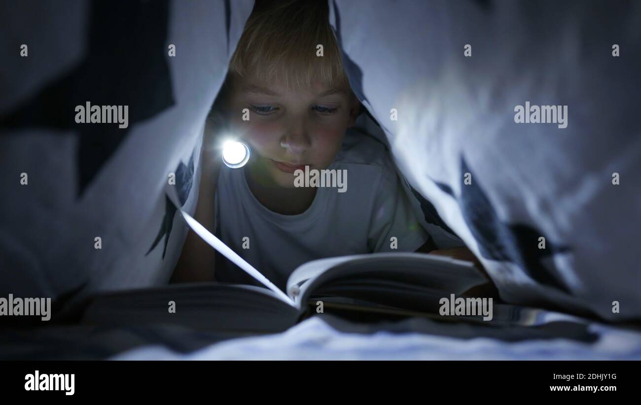 Ein kleiner Junge liest nachts ein Buch mit einer Taschenlampe unter den Deckblättern. Stockfoto