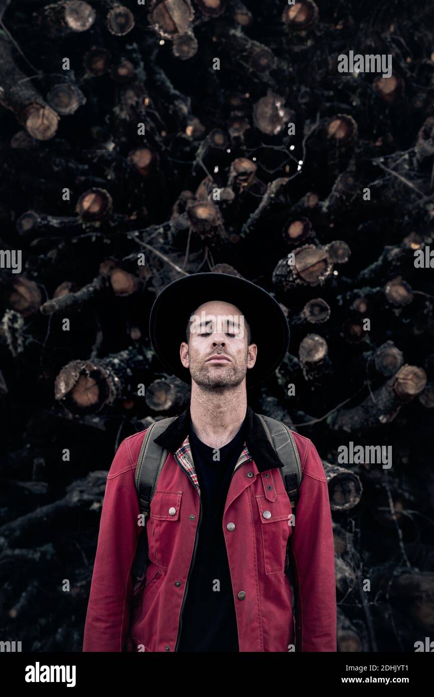 Beschaulicher erwachsener Mann mit Hut und rotem Hemd, der die Augen schließt Gegen Haufen von Baumstämmen in der Natur Stockfoto