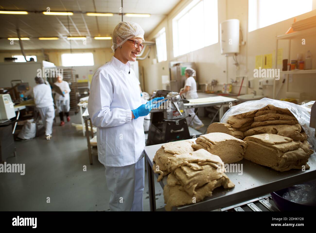 Junge fröhliche Arbeiterin in sterilen Tüchern hält eine Tablette und überprüft die Qualität der Cookie Teig Mischung vor der weiteren Verarbeitung. Stockfoto