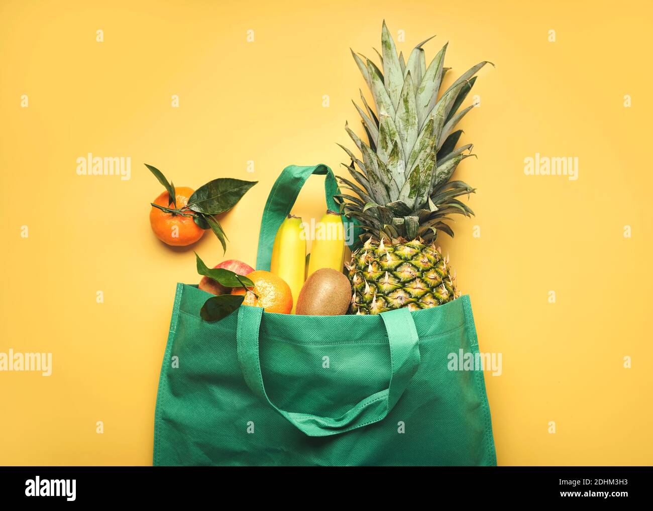 Grüne Einkaufstasche mit verschiedenen Früchten, Ananas, Bananen, Orangen, Kiwis und Äpfeln auf gelbem Hintergrund Stockfoto