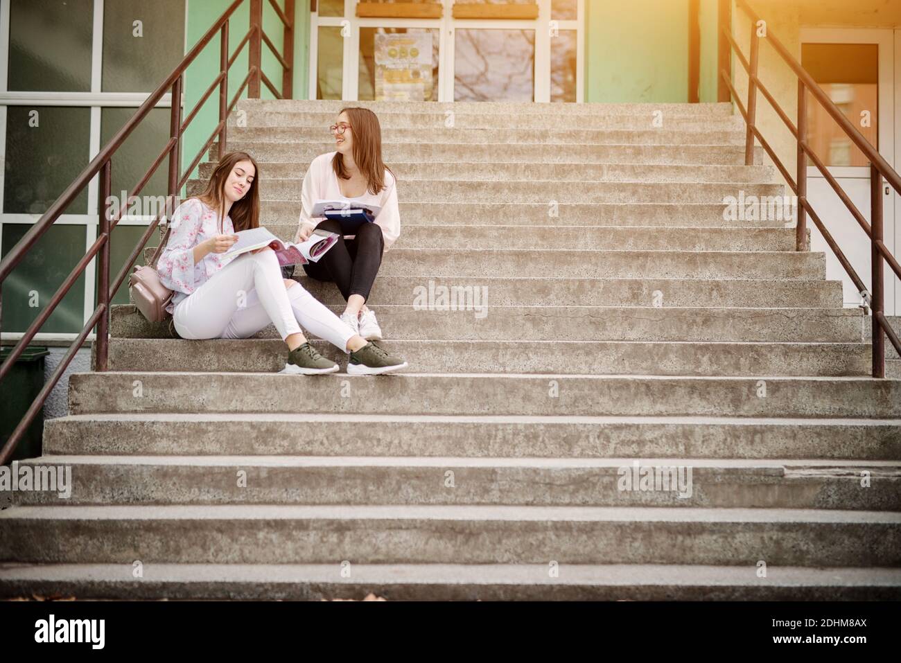 Zwei lächelnde hübsche High School Mädchen sitzen auf Treppen außerhalb der Schule und machen Hausaufgaben zusammen. Stockfoto