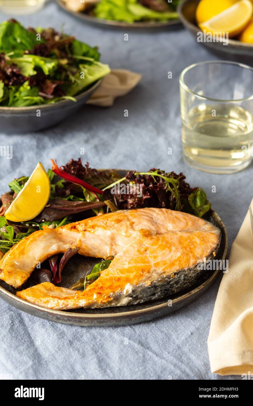 Gebratenes Lachssteak mit Salat auf einem Teller, Weißwein im Glas, leckeres herzhaftes Abendessen oder Mittagessen Stockfoto