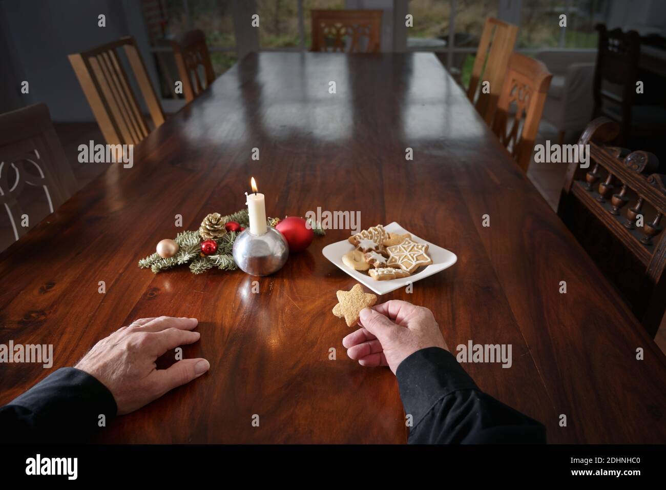 Hände eines einsamen alten Mannes mit einem Cookie sitzt an einem großen Familientisch mit Kerze, Weihnachtsdekoration und leeren Stühlen, Urlaub während der Coronavi Stockfoto