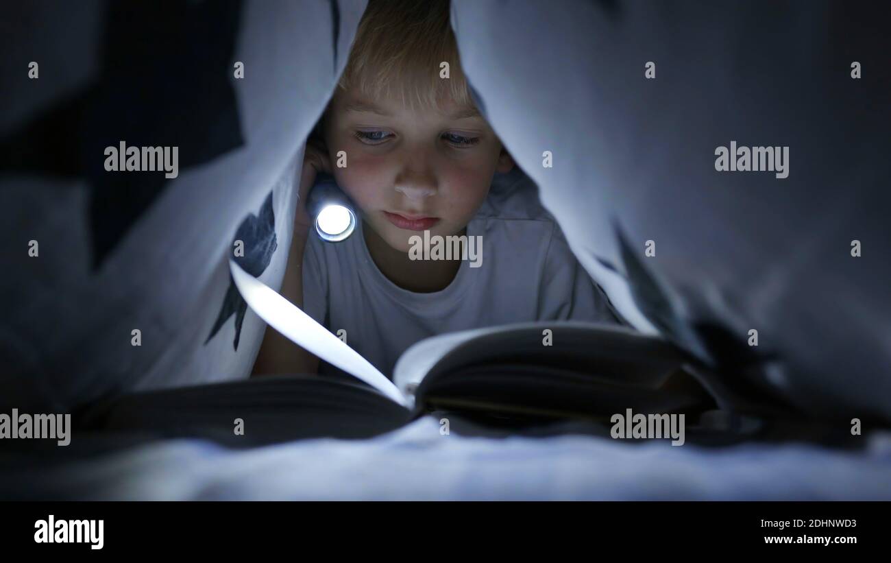 Ein kleiner Junge liest nachts ein Buch mit einer Taschenlampe unter den Deckblättern. Stockfoto