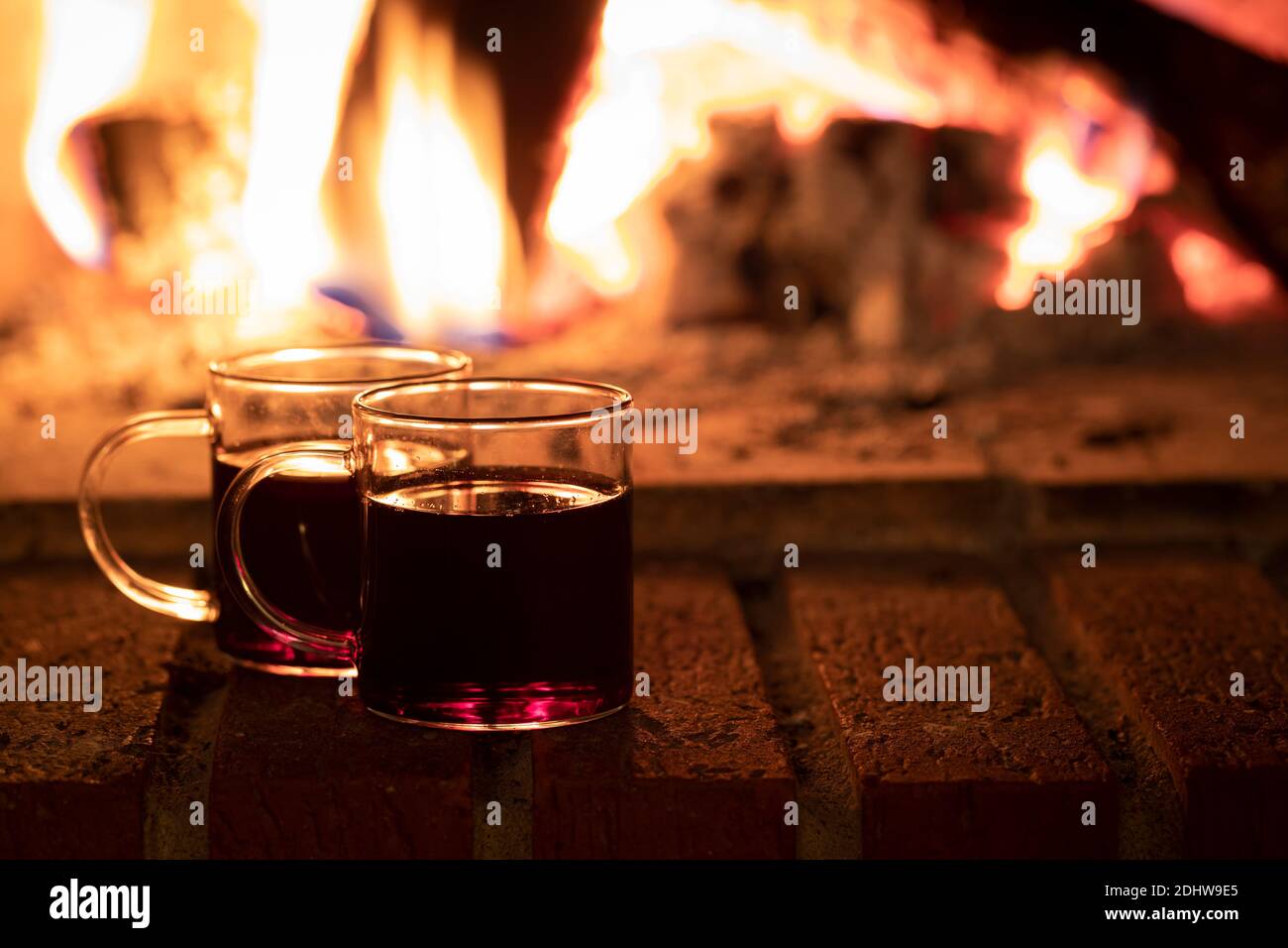 Zwei Tassen Wein oder Glühwein am Feuer. Die Tassen stehen an einem Ziegelkamin mit einem Feuer im Hintergrund, und es gibt Kopierplatz in der Stockfoto