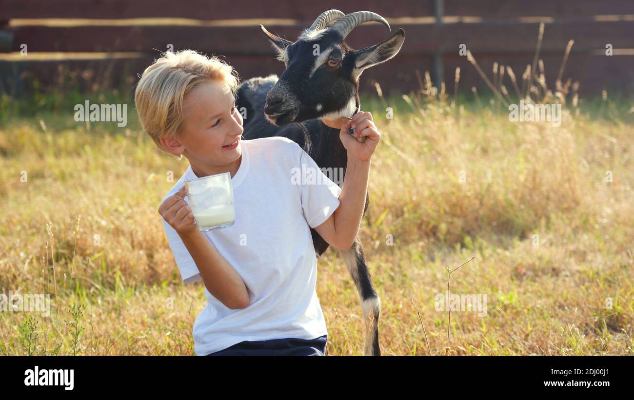 Ein Junge trinkt Ziegenmilch aus einem Becher neben seiner Ziege. Stockfoto