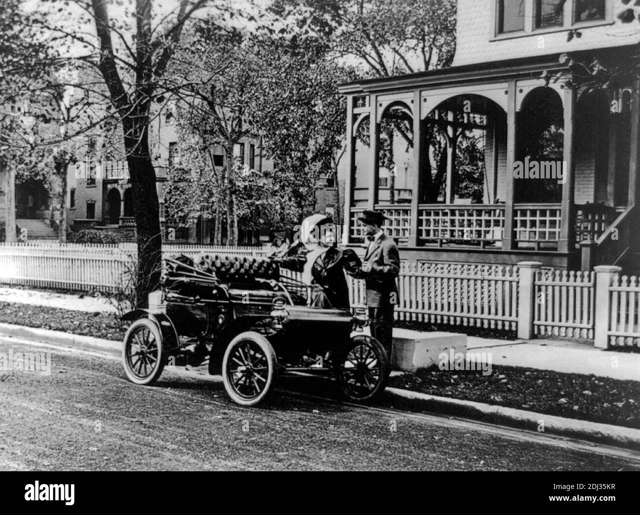 Oldsmobile -Foto zeigt Mann hilft Frau in Auto mit top down geparkt auf der baumgesäumten Straße vor dem Haus, um 1907 Stockfoto