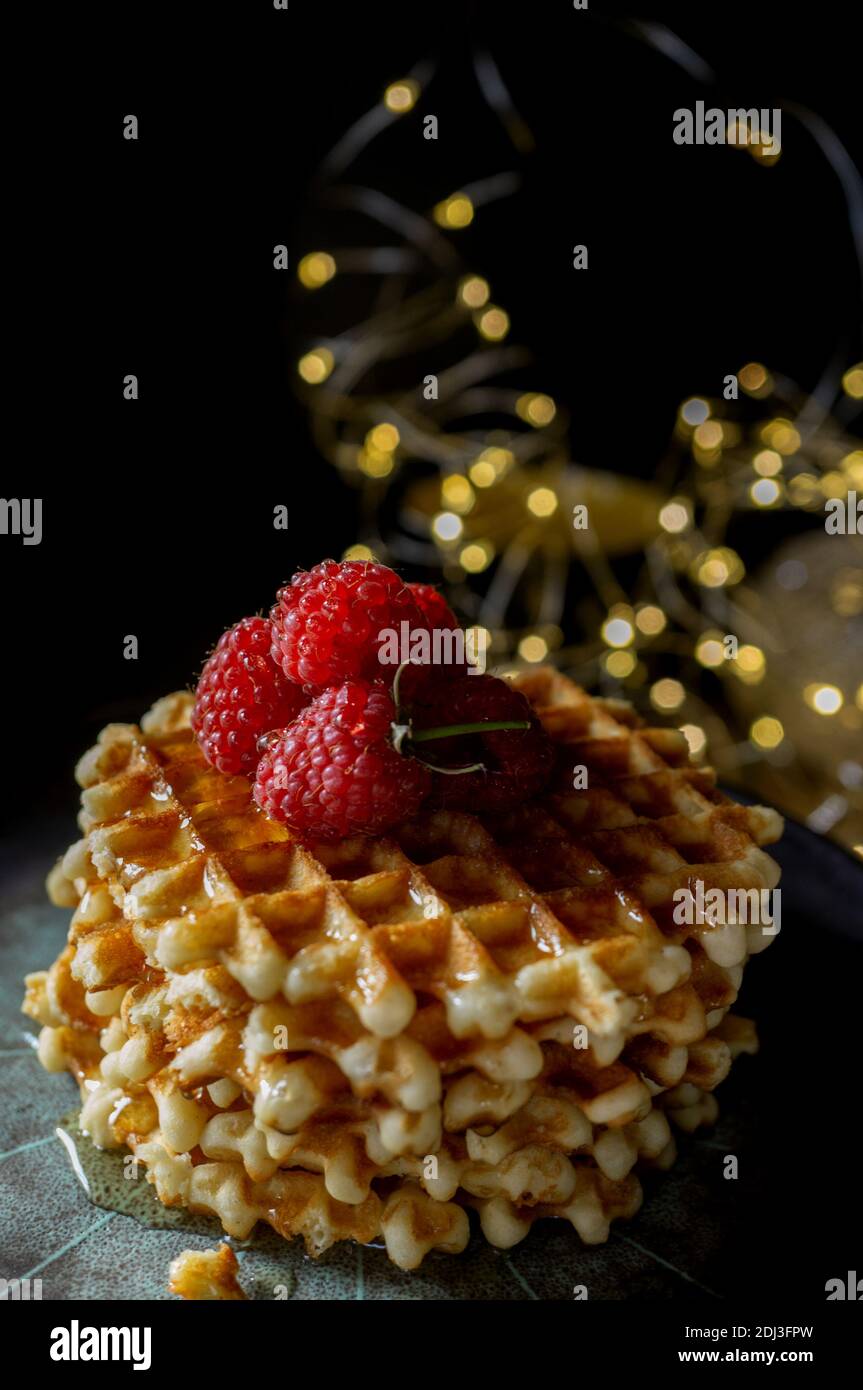 Wiener Waffeln mit Himbeeren und Honig auf dem Weihnachtstisch mit Christbaumzweigen und Girlande-Lichtern im Hintergrund. Stockfoto