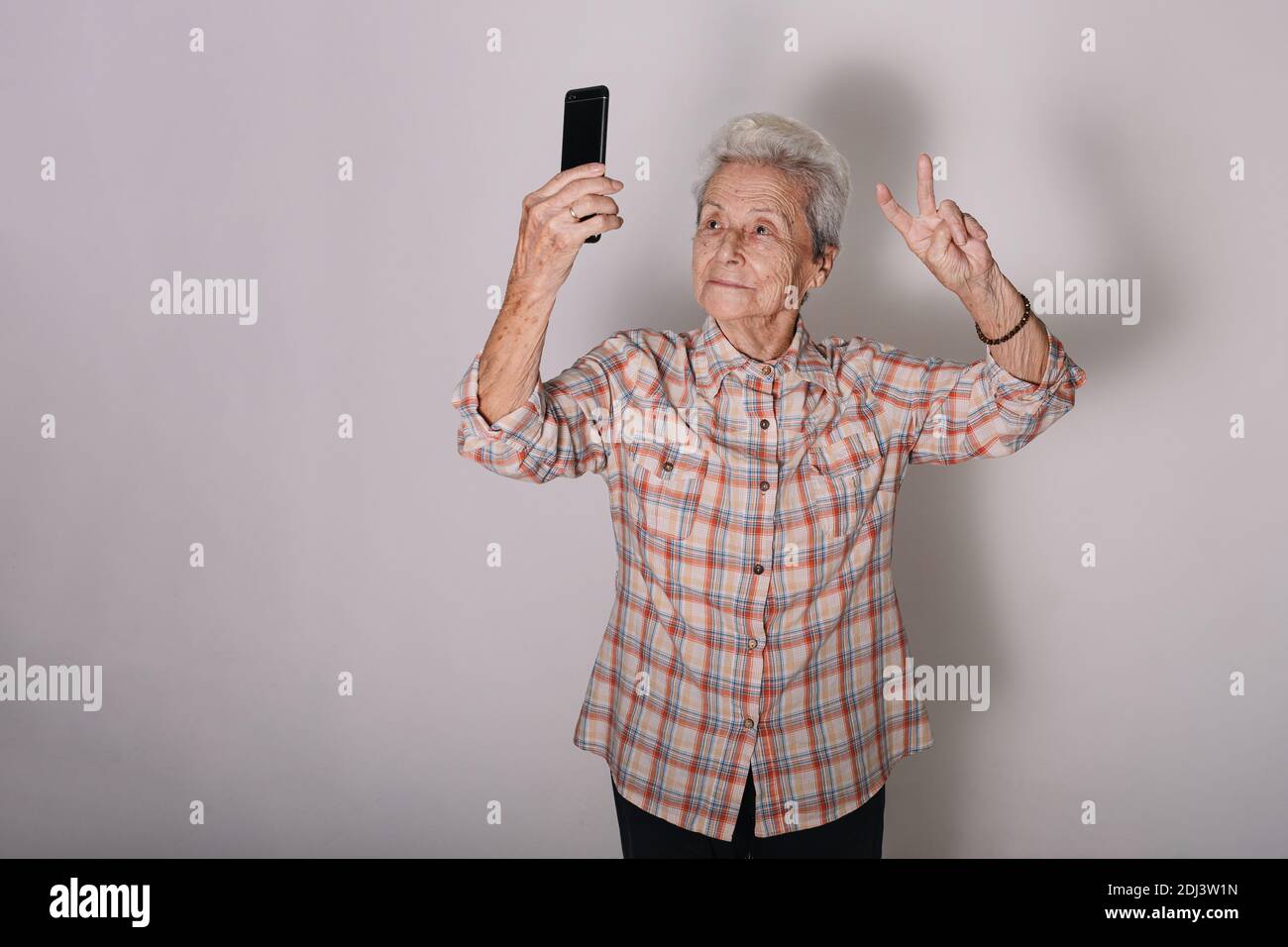 Alte Frau macht ein Selfie mit einem intelligenten, glücklichen Telefon und das Symbol des Friedens oder des Sieges in der Hand. Technologisches Konzept. Aktives Alterskonzept. Gra Stockfoto