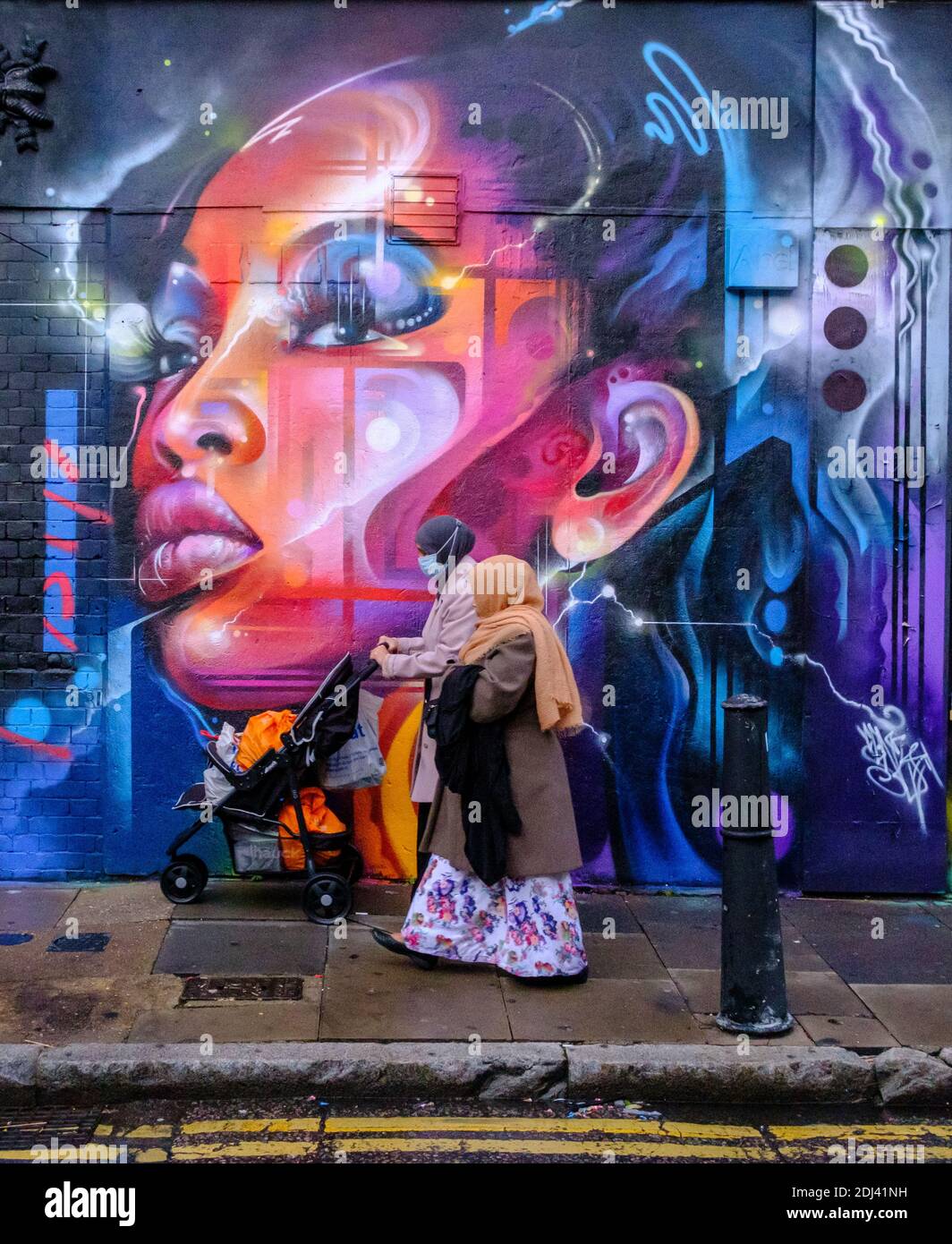 Zwei Frauen in Kopftüchern mit Kinderwagen laufen vor dem Street Art Wandbild von Mr. Cenz in der Bacon Street & Brick Lane, Shoreditch, East London, England Stockfoto