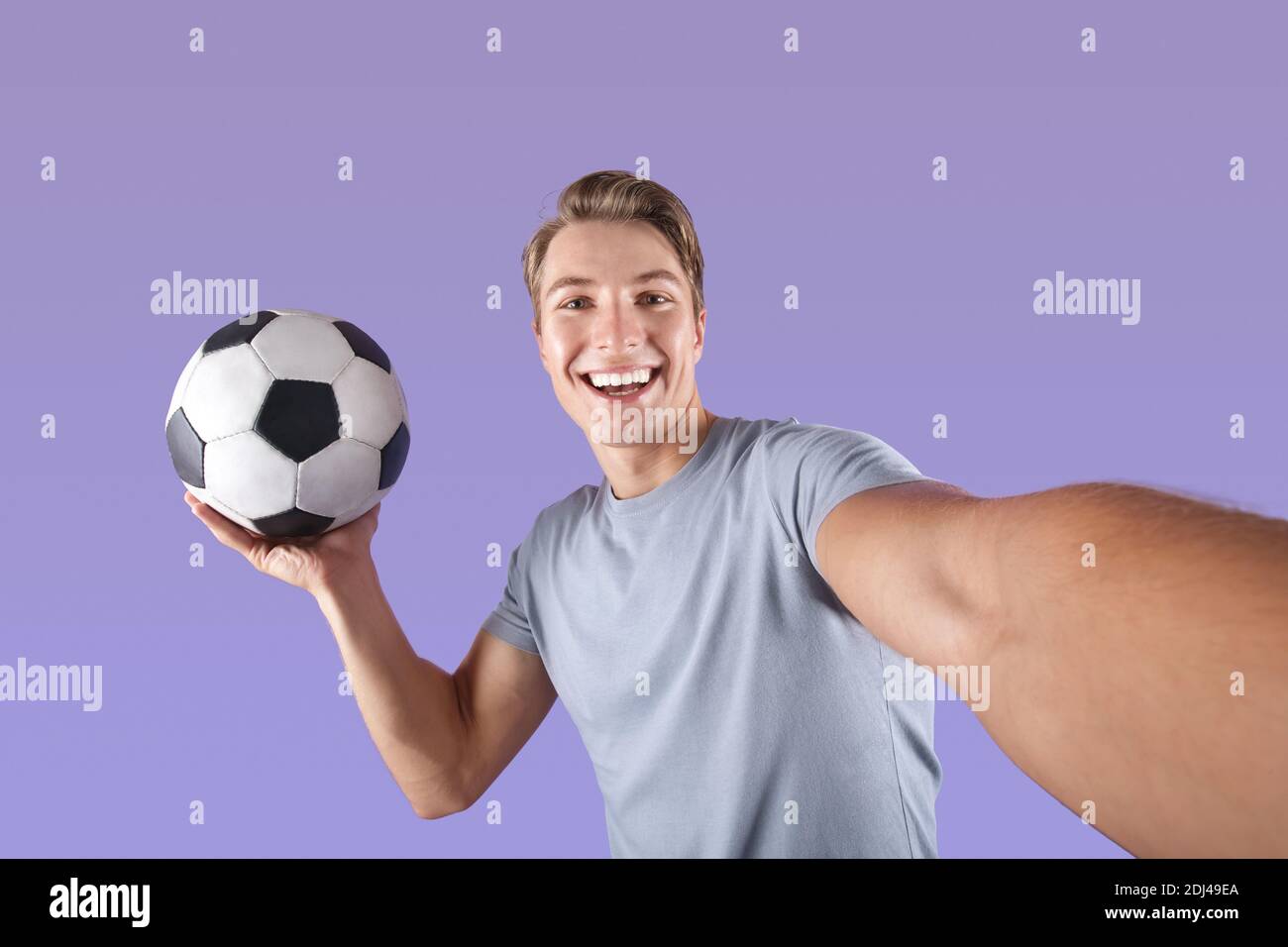 Lächelnder junger Mann mit Fußball nimmt Selfie auf seine Mobiltelefon über violettem Studiohintergrund Stockfoto