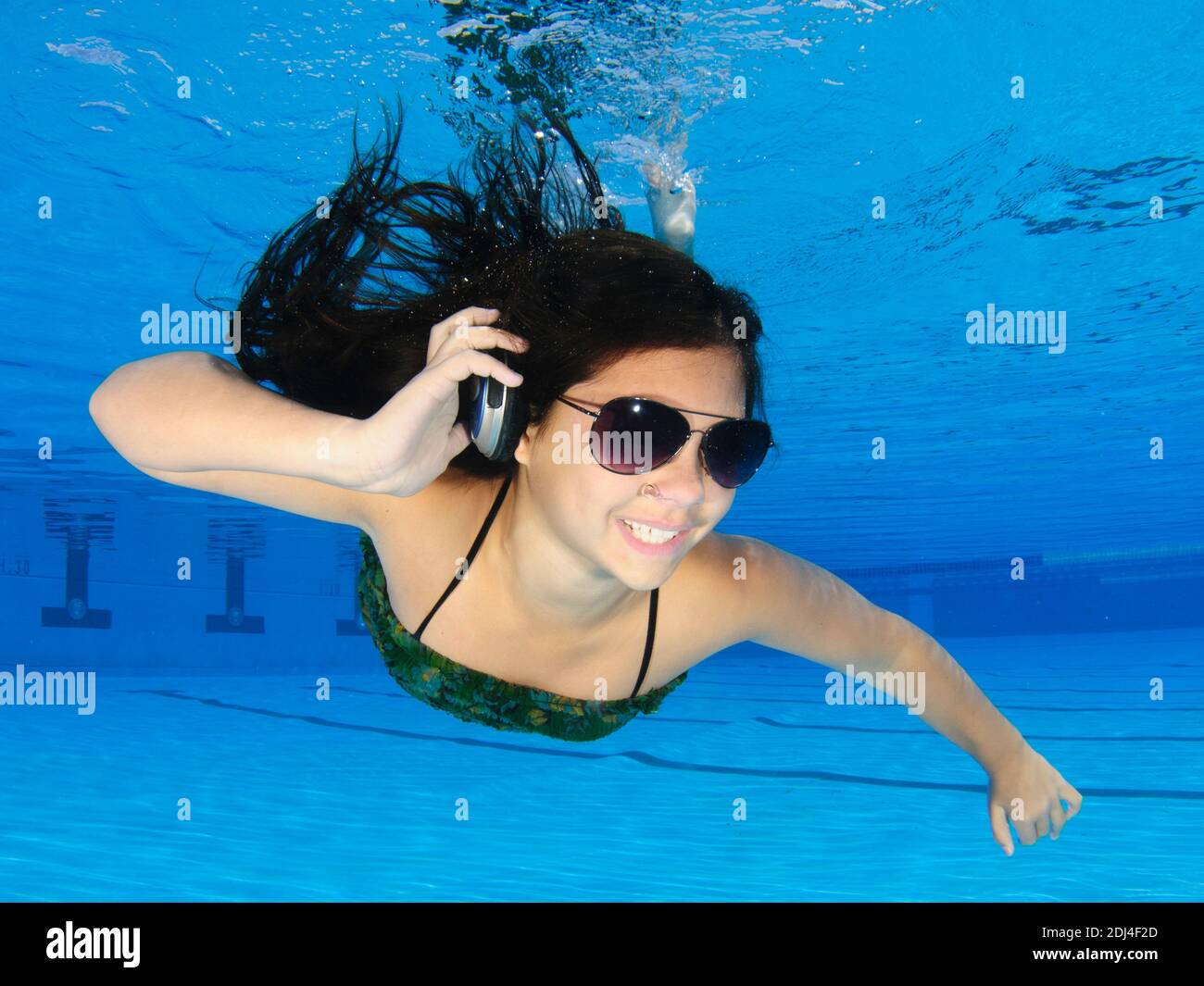 Eine gekleidete 12-jährige Teenager-Frau, die unter Wasser in einem Schwimmbad mit Kopfhörern und Sonnenbrillen tauchen kann. Modellversion verfügbar Stockfoto