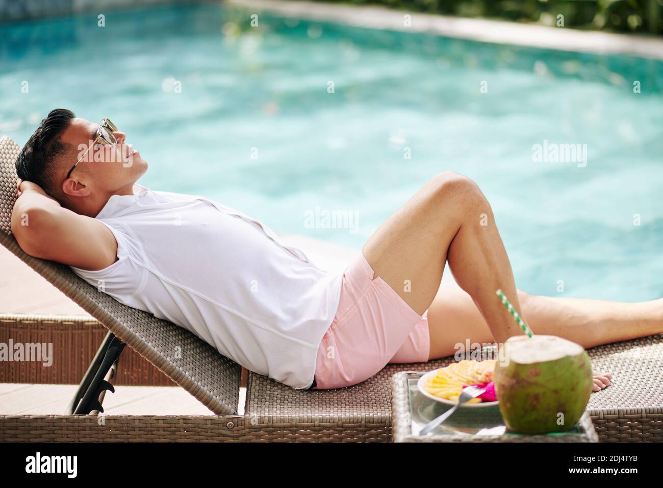 Fröhlicher junger Mann in Sonnenbrille, der beim Schwimmen auf der Chaiselongue ruht Pool mit Kokosnusscocktail und Obstteller auf dem Tisch In der Nähe Stockfoto