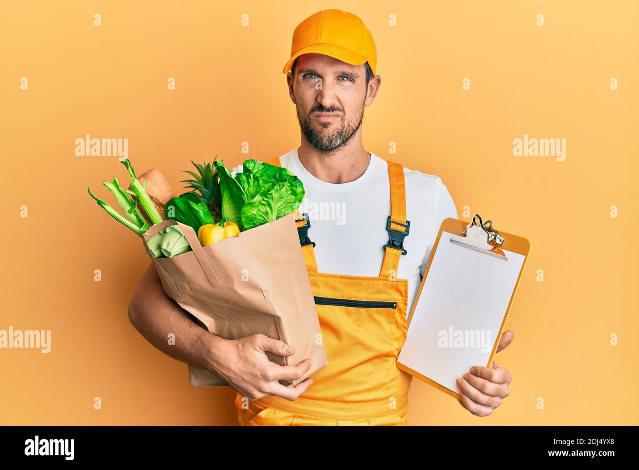Junger schöner Mann trägt Kurieruniform mit Lebensmitteln und Zwischenablage ahnungslos und verwirrt Ausdruck. Zweifel Konzept. Stockfoto