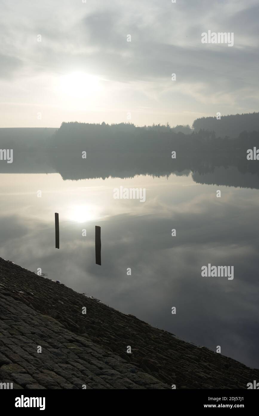 Zwei Pfosten umrahmen die Wintersonne, wie sie im ruhigen Wasser eines südlichen yorkshire Reservoirs reflektiert wird. Wald Hintergrund und Staumauer Vordergrund. Stockfoto