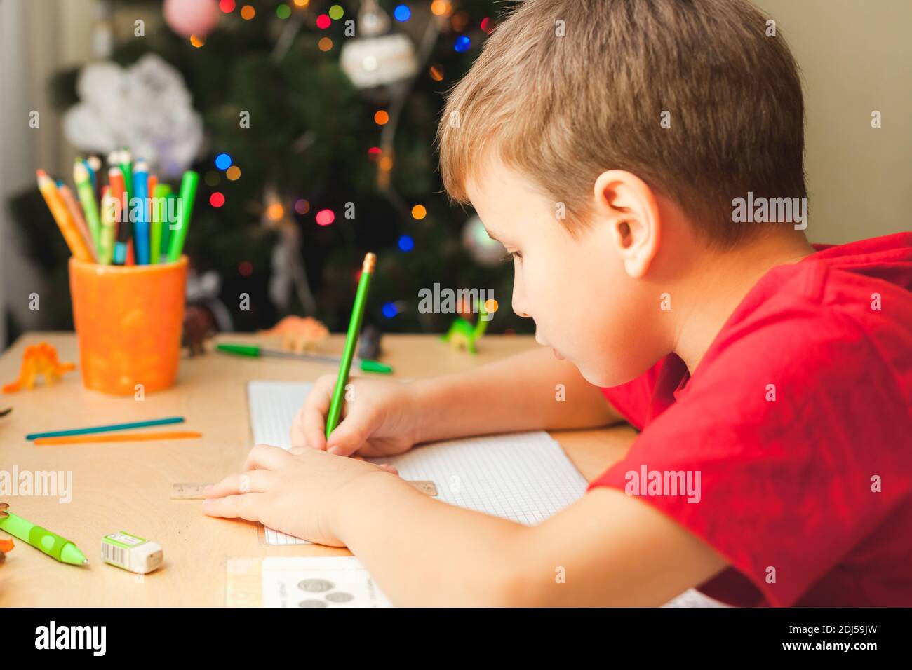 7 Jahre alter Junge, der am Schreibtisch sitzt und Hausaufgaben macht. Kind schreibt Notizen im Notizbuch, schwierige Hausaufgaben, Kind konzentriert sich auf Beispiele. Geschmückter Weihnachtsbaum im Hintergrund Stockfoto