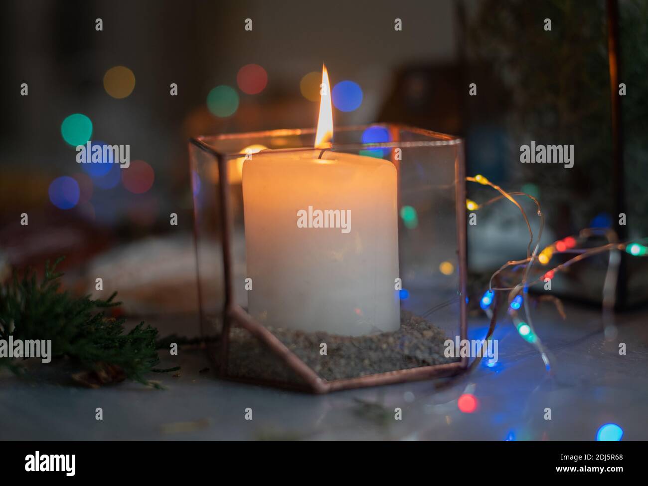 Eine brennende Kerze in einem Glashalter in einer stimmungsvollen gemütlichen Atmosphäre zu Hause in der Nacht durch das Licht einer Girlande. Urlaub, Weihnachten und Neujahr Stockfoto